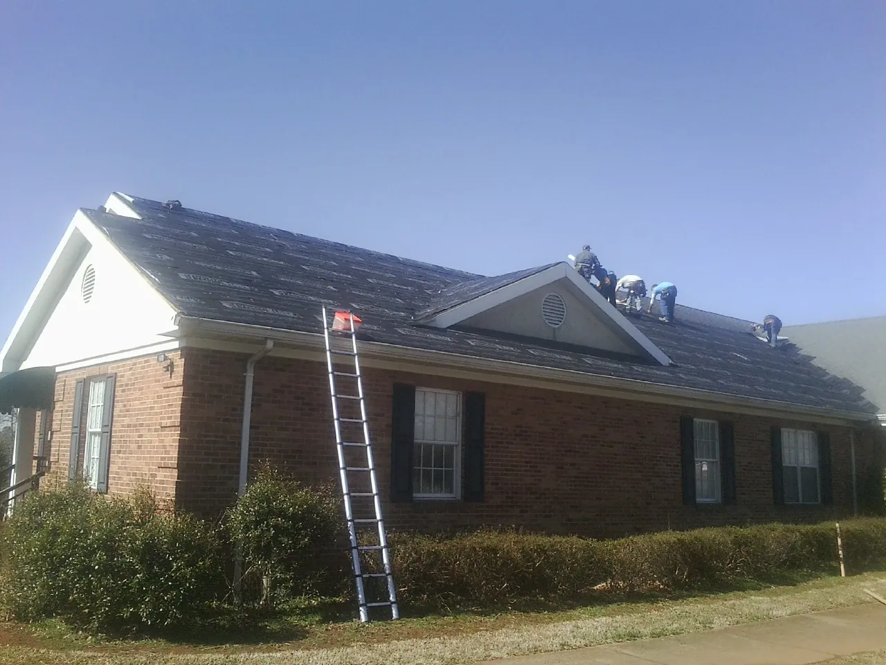 Crew tearing shingles off a brick ranch roof with a ladder set — Emergency Roof Repair work by our crew in Aragon