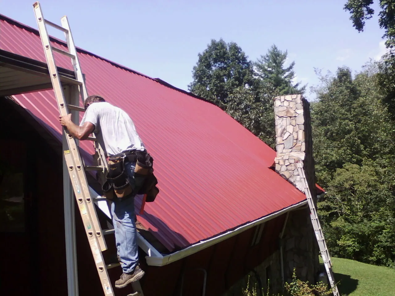 Roofer on a ladder beside a red metal roof and stone chimney — Soffit & Fascia Repair by Roofing Brothers in Aragon